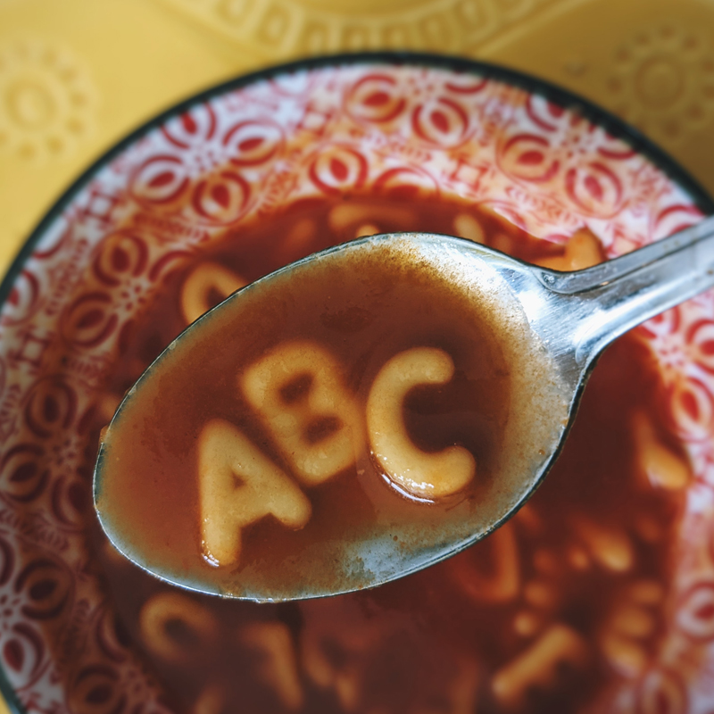 ABC pasta in a spoonful of red tomato soup held above a bowl of soup.